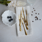 Six gold-toned flatware forks arranged on a folded cream linen napkin, styled on a light stone surface with a small white bowl of blueberries, scattered coffee beans, flowers, and a glass at the top.