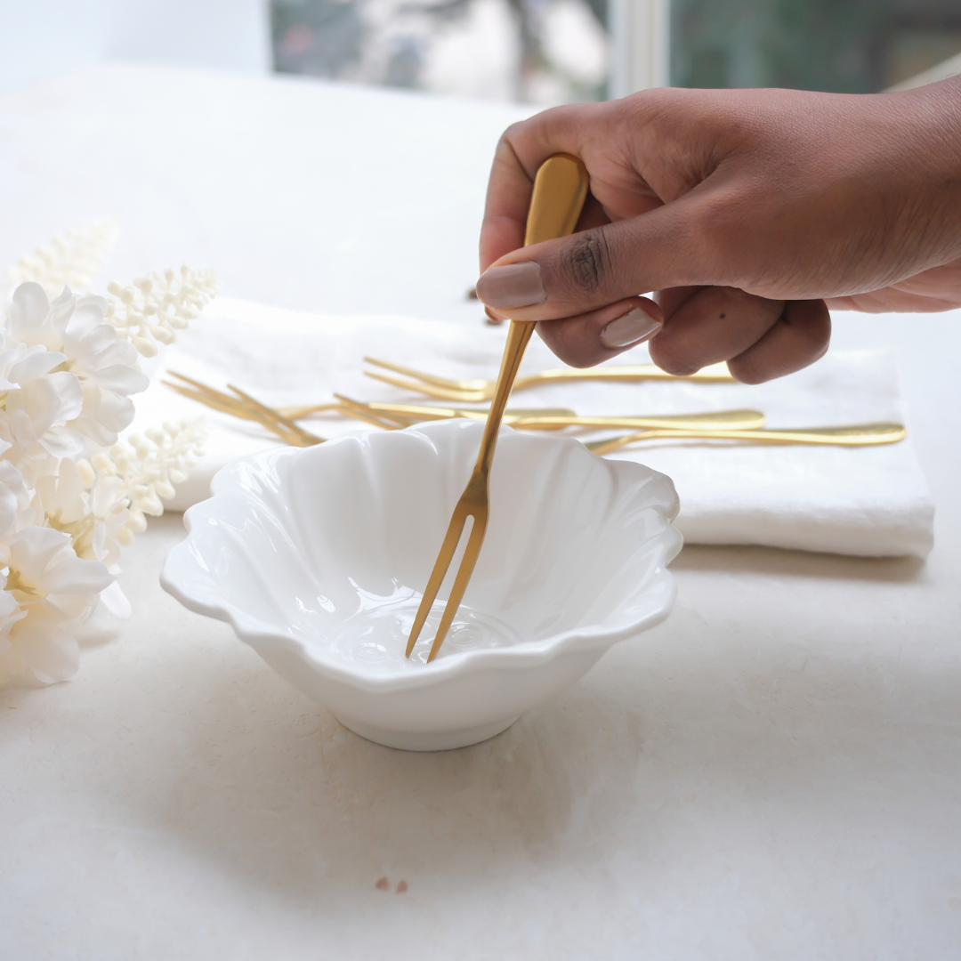A hand holding a gold-toned serving fork over a small white ceramic bowl on a light tabletop, with additional gold utensils and white flowers in the background.