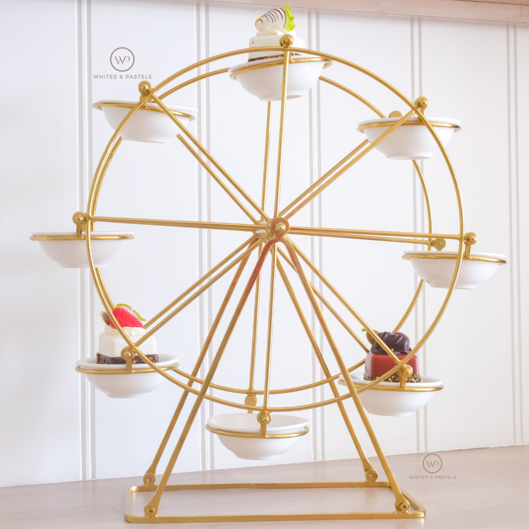 A gold metal Ferris wheel stand with six white ceramic cups or bowls attached at different wheel positions, displayed on a tabletop against a white paneled wall. Small dessert items are placed in some of the cups.