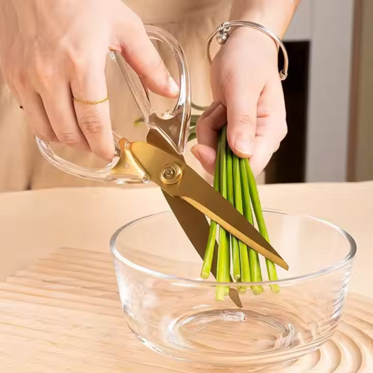 Person using gold scissors to cut green vegetables over a clear glass bowl on a wooden surface.