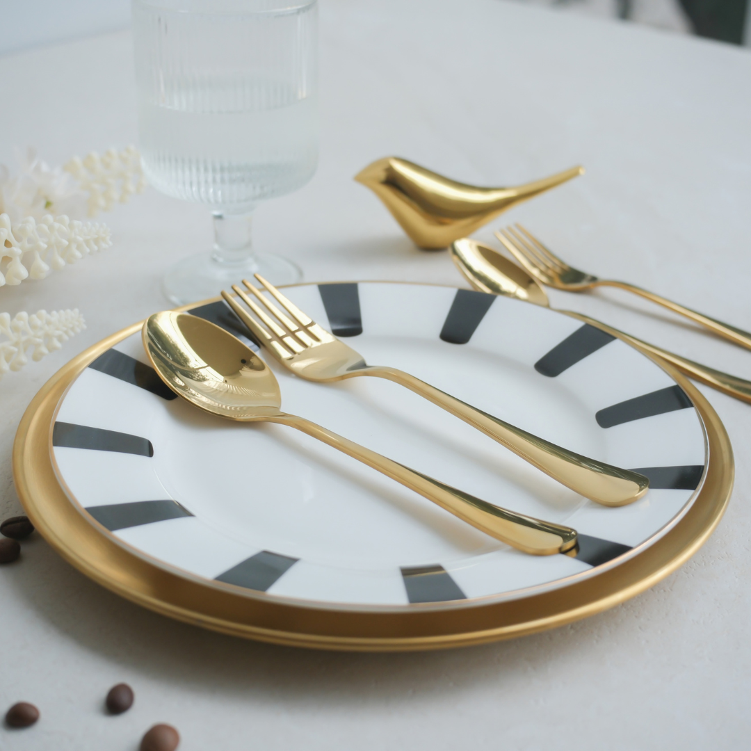 Gold flatware set including a spoon, fork, and bird-shaped utensil, arranged on a white plate with black stripes and a gold rim, on a light tabletop.
