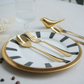 Gold flatware set including a spoon, fork, and bird-shaped utensil, arranged on a white plate with black stripes and a gold rim, on a light tabletop.