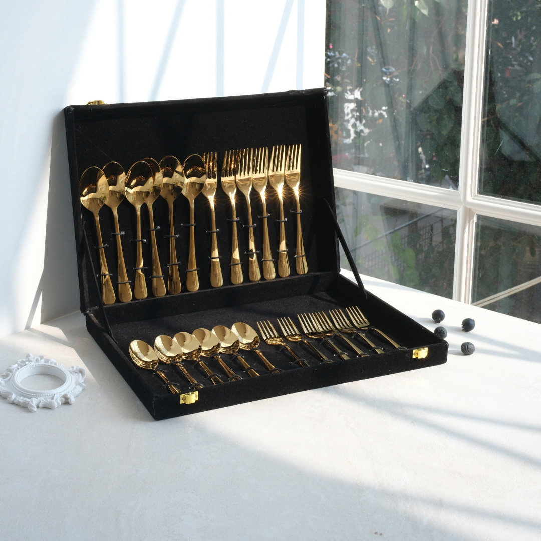 Gold flatware set with matching black velvet case, displayed open on a white table near a window.