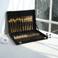 Gold flatware set with matching black velvet case, displayed open on a white table near a window.