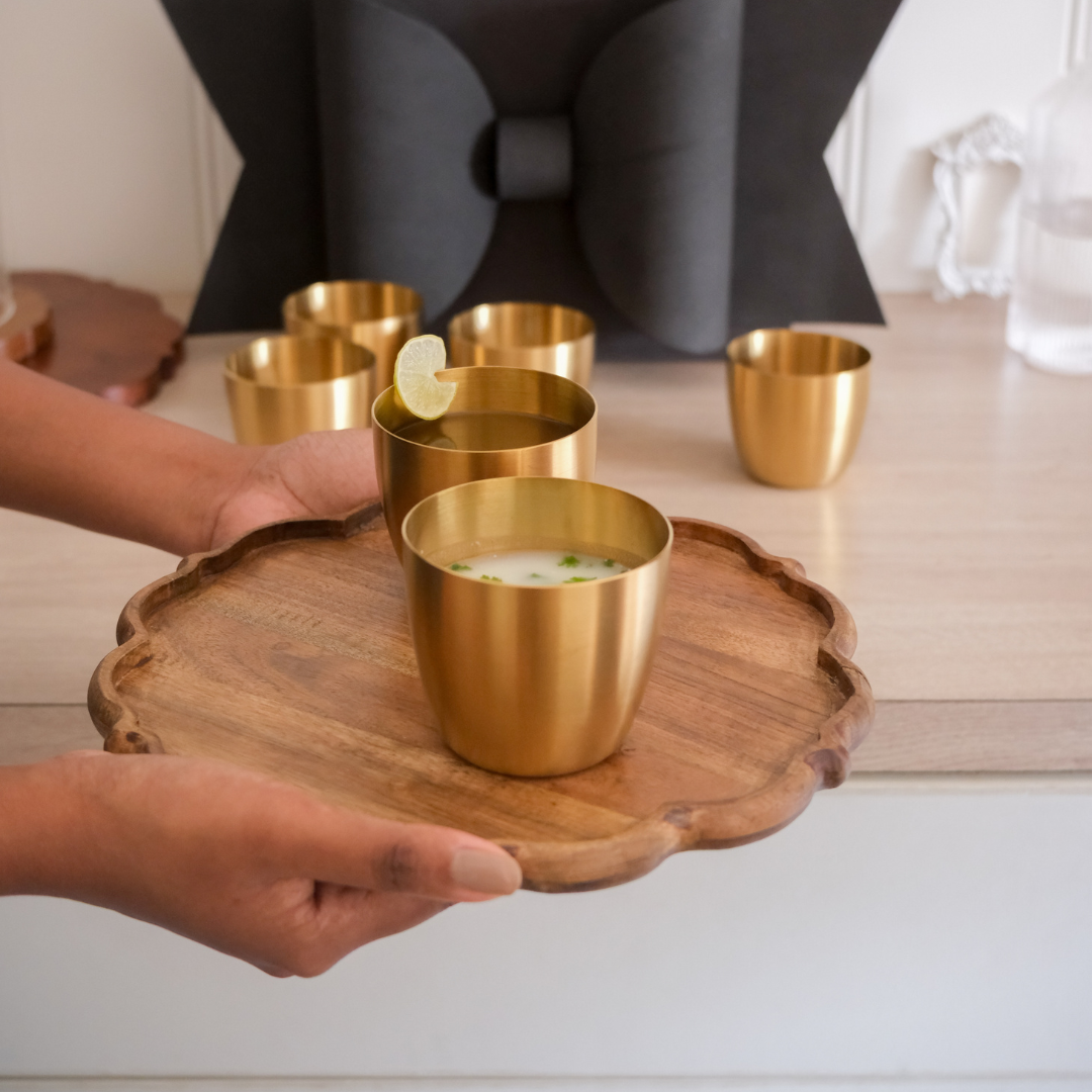 A set of small gold cups tumbler arranged on a scalloped wooden tray, with a person's hands holding the tray in a bright indoor setting.
