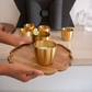 A set of small gold cups tumbler arranged on a scalloped wooden tray, with a person's hands holding the tray in a bright indoor setting.