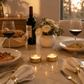 A warmly lit dining table set for two, featuring a bottle of red wine, two filled wine glasses, plates of food, a vase of white flowers, and two lit gold candle holders on a marble surface.