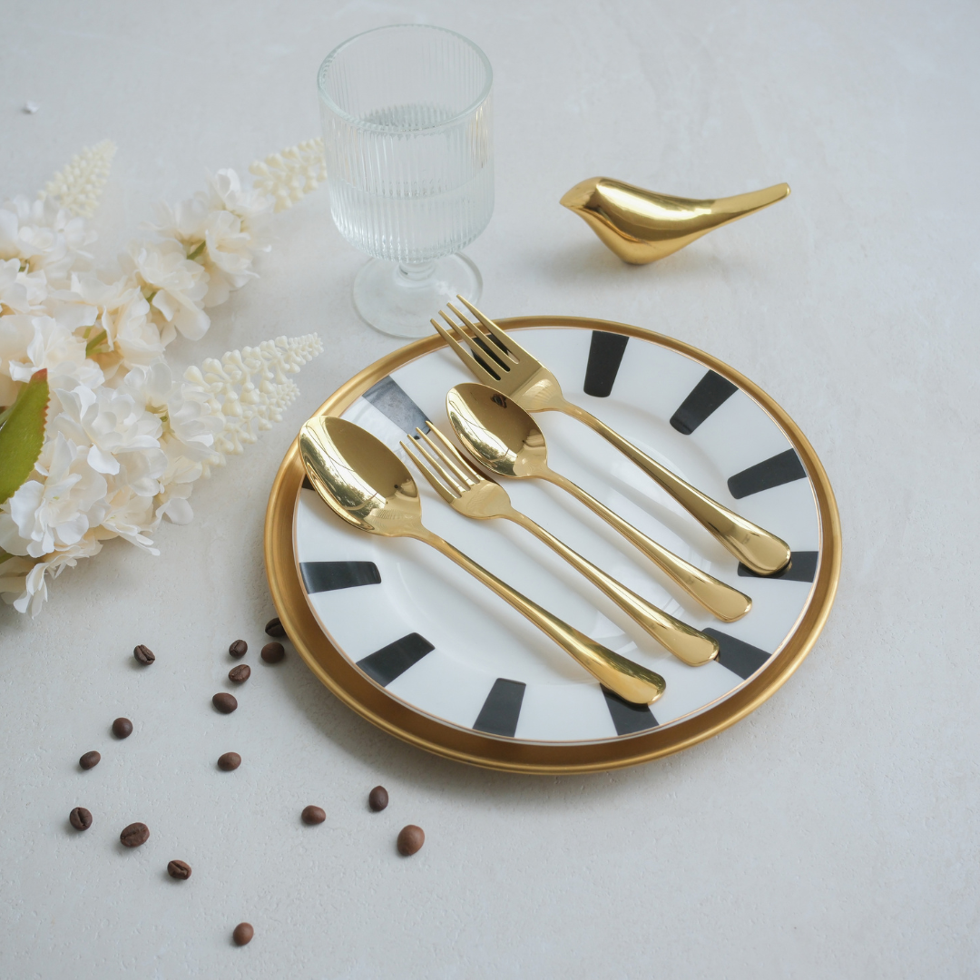 Gold flatware set with spoons and forks on a white and black patterned plate, styled with a glass, decorative flowers, coffee beans, and a gold bird-shaped object on a light gray surface.