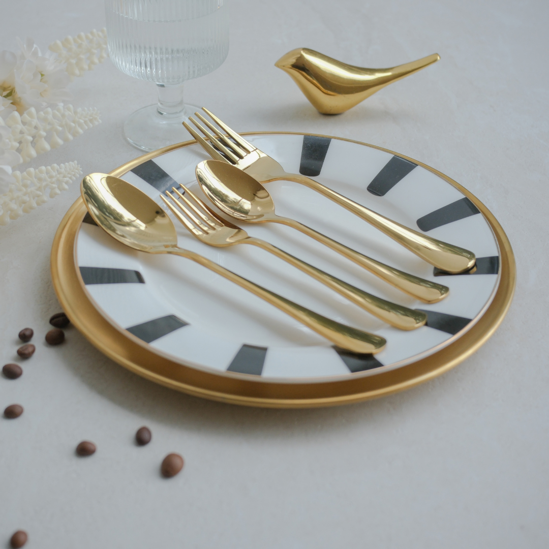 A set of four gold-toned flatware pieces, including two forks and two spoons, arranged on a white ceramic plate with black geometric stripes and a gold rim, styled on a light gray surface with coffee beans, a glass, and a small gold bird figurine.