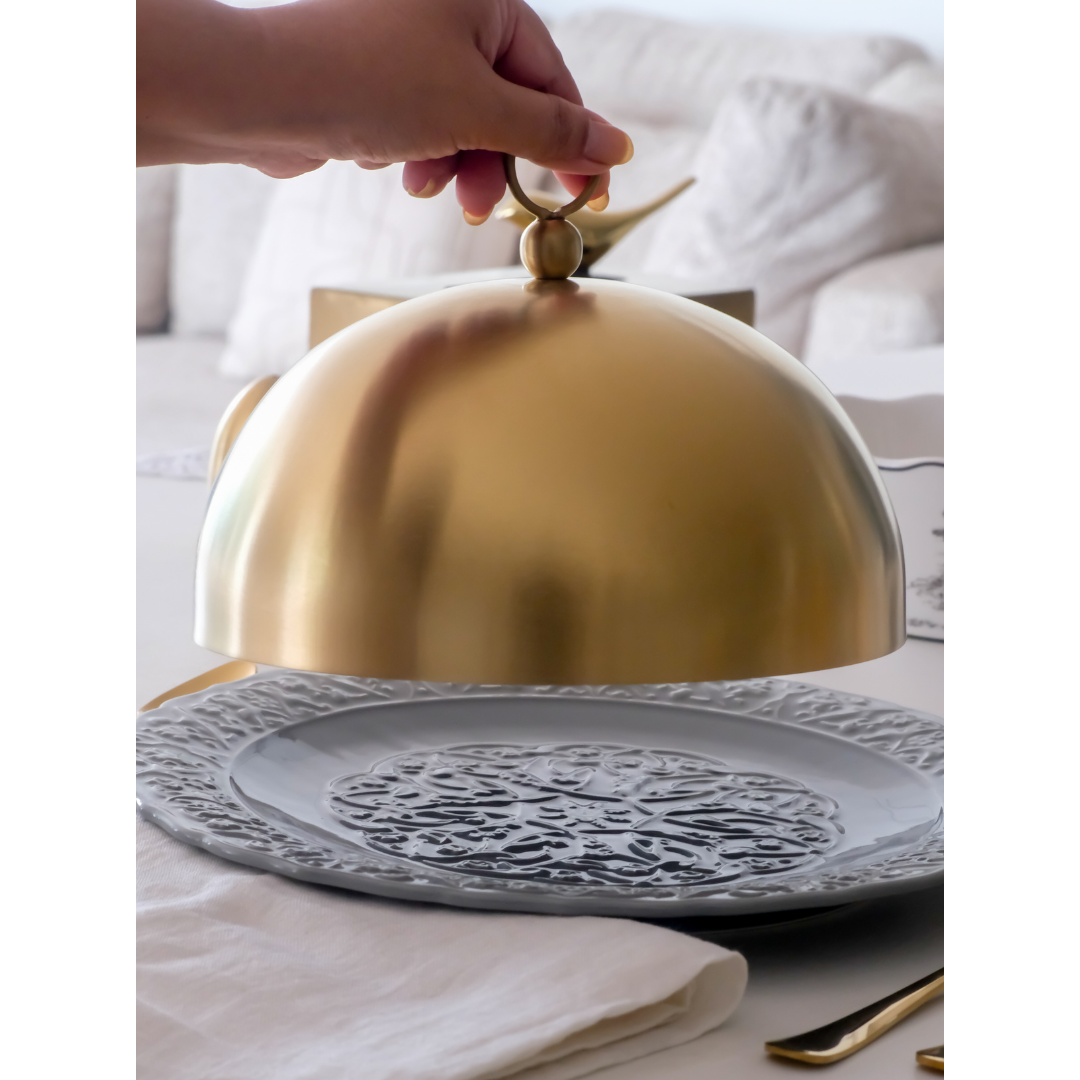 Gold cloche on a decorative plate with a hand lifting the lid, set against a neutral background.