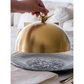 Gold cloche on a decorative plate with a hand lifting the lid, set against a neutral background.