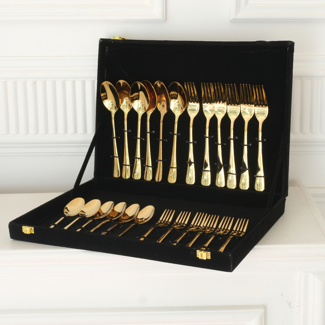 A gold-plated cutlery set with forks, spoons, and knives displayed inside an open black velvet presentation case on a white mantelpiece.