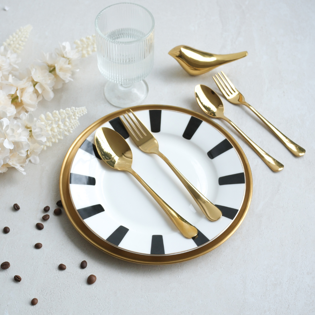 Gold flatware set with a bird-shaped spoon rest, shown on a white plate with black stripes and a gold rim, styled with a glass of water, white flowers, and coffee beans on a light gray surface.