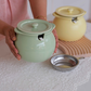 A person holds a pale mint green ceramic jar with a lid and small side spout, while a matching pale yellow jar sits in the background on a wooden board. A small stainless steel bowl is placed in front on a white lace tablecloth.
