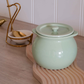 A glossy pale green ceramic pot with a matching lid sits on a wavy wooden board on a light wood tabletop. A small white bowl with a gold spoon in a gold stand is visible in the background against a white paneled wall.