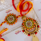 A pair of traditional Indian Rakhi Set displayed on a round gold-colored plate. This Rakhi for Bhai and Bhabi features intricate floral mandala designs with green, orange, and red stones, white pearl-like beads, and red tassels, along with a gold leaf charm.