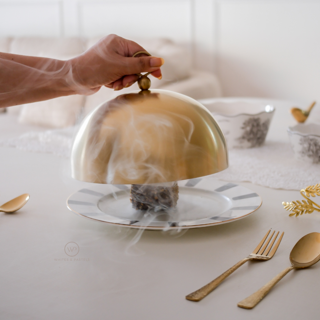 Hand lifting a gold cloche over a dish on a table with gold cutlery.