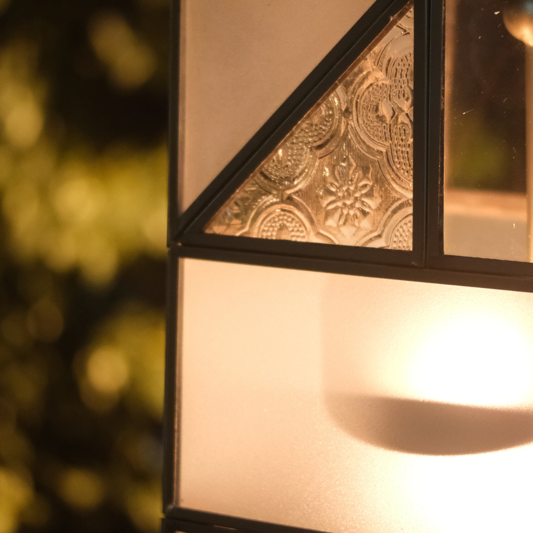 Close-up of a decorative outdoor light fixture with black metal framing, a triangular section of embossed textured glass, and a warm glowing frosted panel.