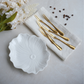 A white ceramic flower-shaped plate with a scalloped rim and embossed center, styled on a light surface with four gold forks on a folded white napkin, white flowers, and a few coffee beans.