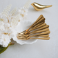 A set of gold stainless steel forks arranged in a small white ceramic dish, styled with white flowers on a light gray surface. A matching gold bird-shaped object is visible in the background.