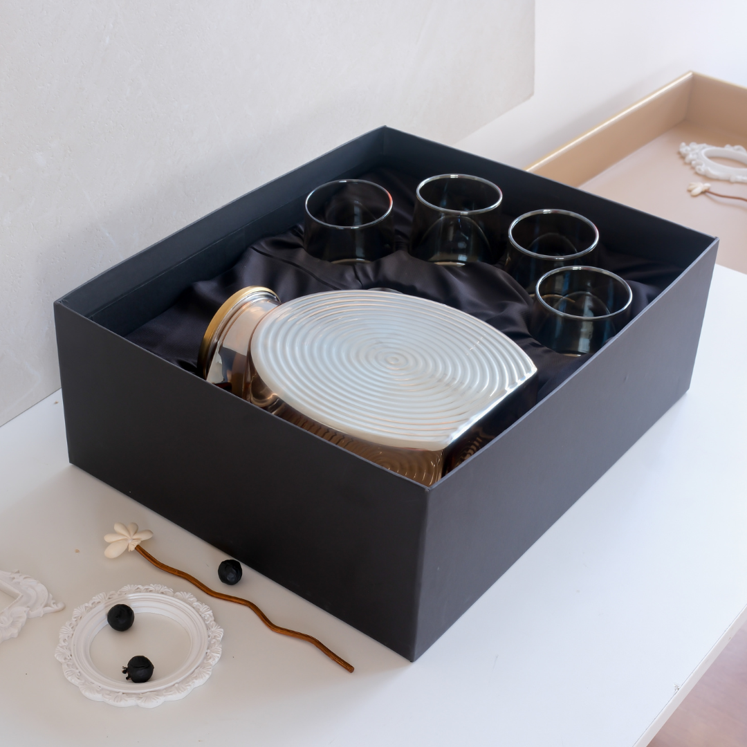 A boxed glassware and decanter set displayed in an open black gift box on a white surface. The set includes a ribbed white decanter with a gold-toned neck, four dark translucent glasses, and black satin lining inside the box.