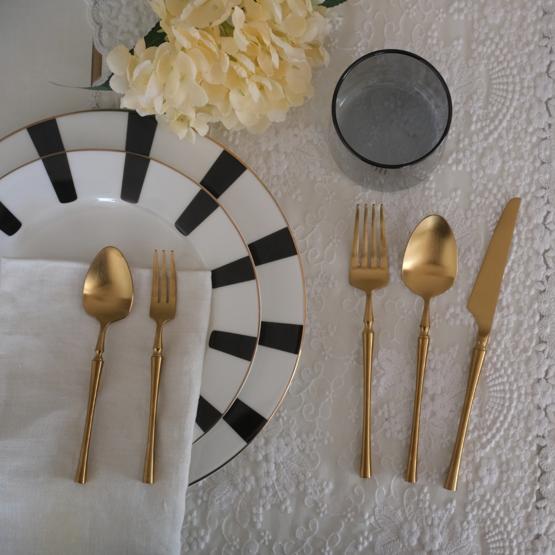 Gold flatware set including a spoon, fork, and knife, arranged on a white lace tablecloth alongside a black and white patterned plate, a folded white napkin, and a glass of water.