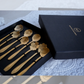A set of gold-toned floral utensils, likely spoons or small tools, presented in an open black gift box next to a closed black box with gold lettering that reads 'Mochi & More'.