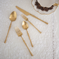 A set of gold-toned flatware, including a knife, two spoons, and two forks, arranged on a white lace placemat with a white teacup and coffee beans in the corner.