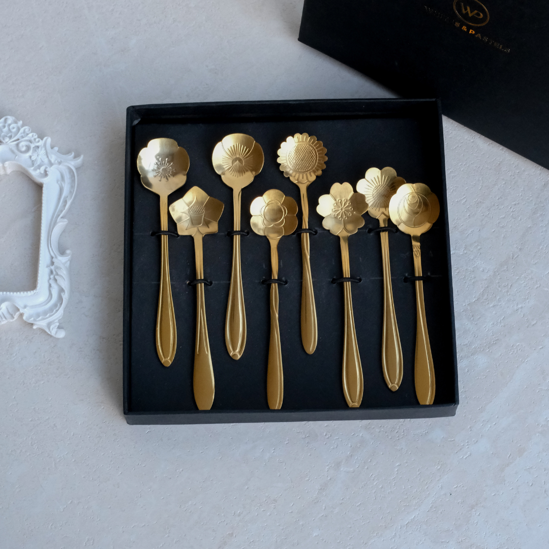 A set of eight gold-toned decorative spoons with unique ornate handles and flower-shaped heads, presented in a black gift box on a light textured surface.