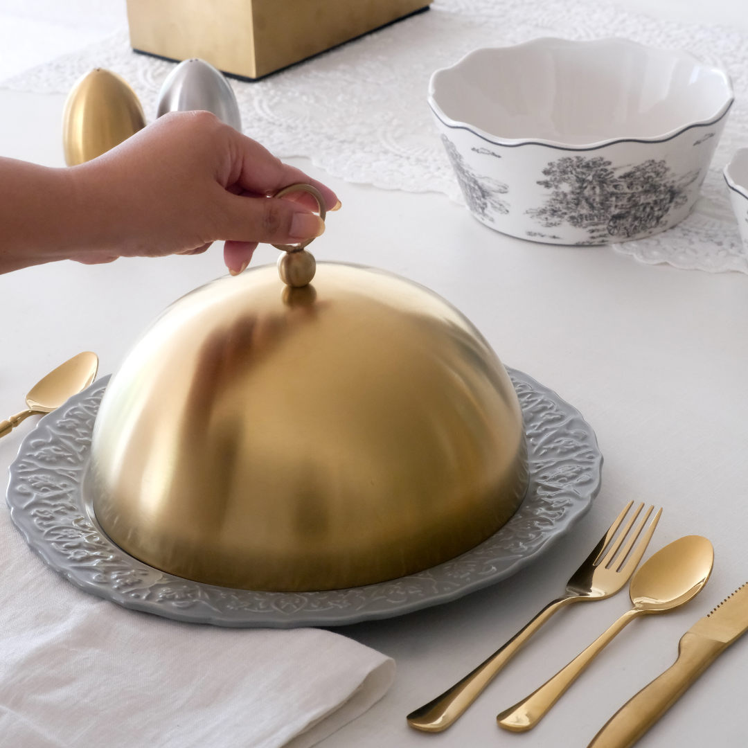 Gold cloche on a gray plate with silverware and a hand placing a gold knob on it.