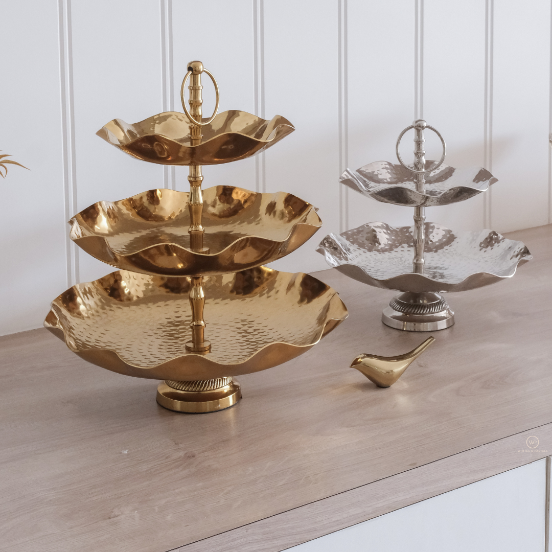 Gold three-tiered serving tray on a wooden surface with a white paneled wall background.