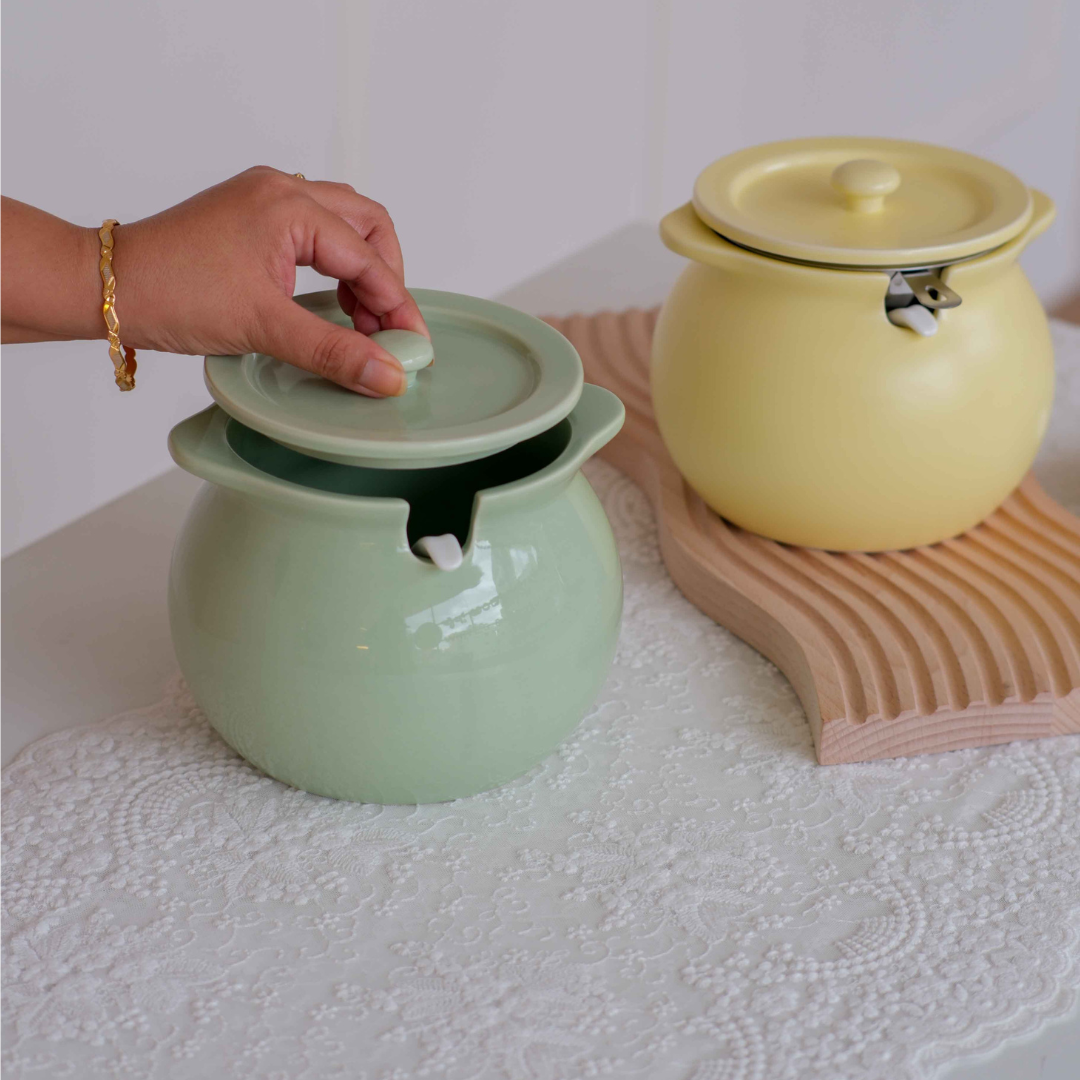 Two ceramic soup pots with lids are shown on a white tablecloth. One pot in the foreground is a pale mint green and is being lifted by a person's hand.