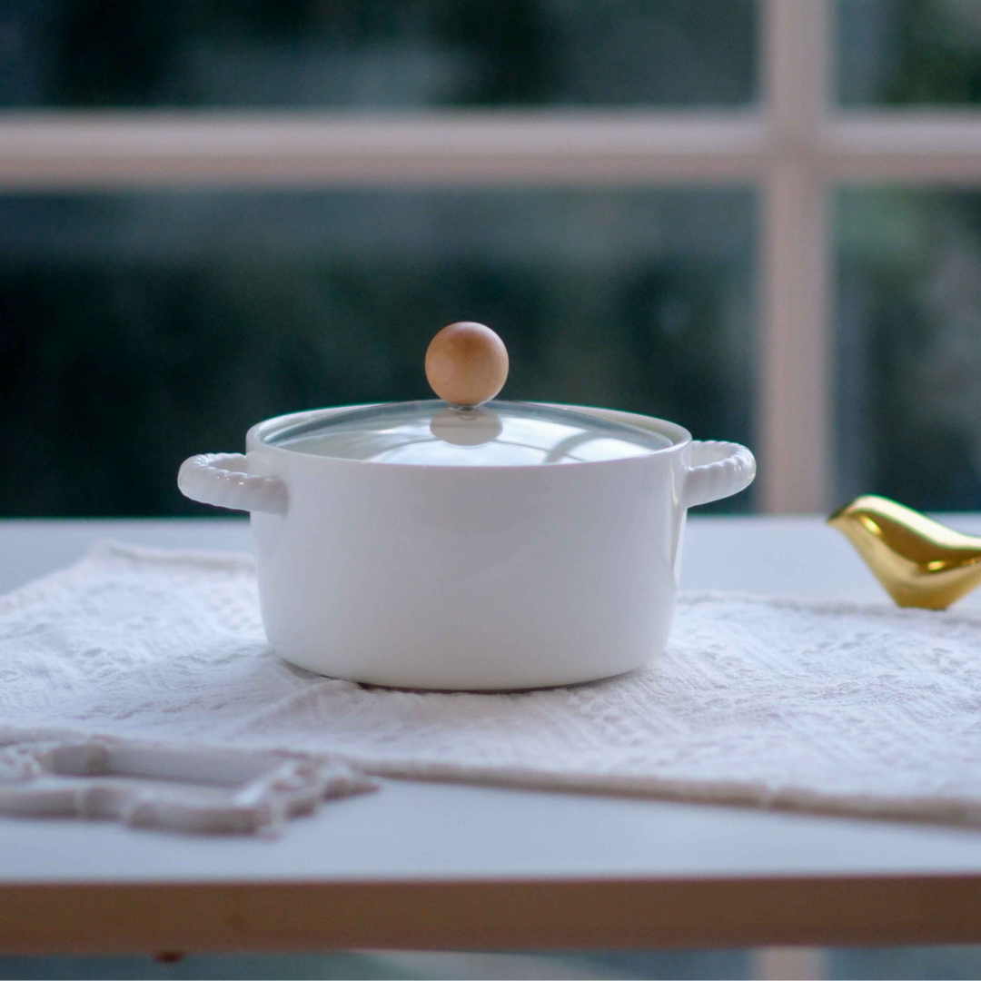 Luxury white ceramic casserole serving bowl with glass lid, rope-style handles and wooden knob displayed on a wooden trivet for contemporary table styling.