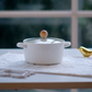 Luxury white ceramic casserole serving bowl with glass lid, rope-style handles and wooden knob displayed on a wooden trivet for contemporary table styling.