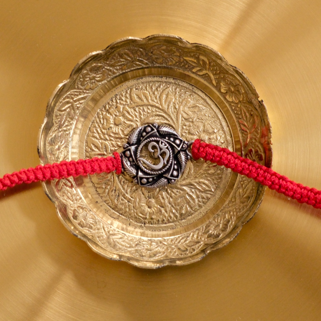Pure Silver Om Rakhi featuring an intricately crafted silver Om motif on a vibrant red braided thread, displayed on an engraved brass plate for Raksha Bandhan.