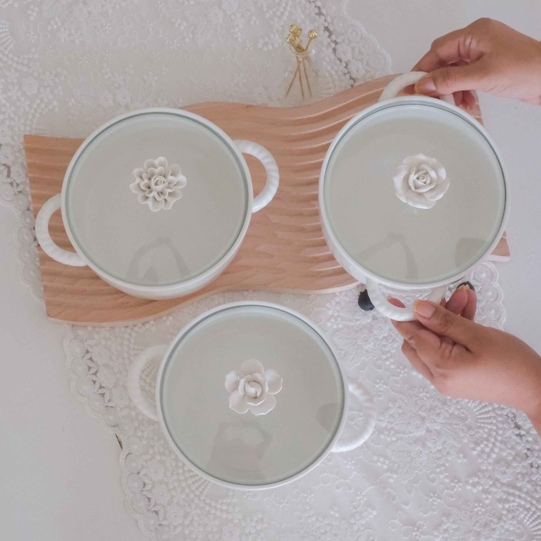 Three white ceramic bowls with matching lids, each featuring a raised white floral design in the center and a glass lid, arranged on a wooden tray over a white lace tablecloth. Two hands are holding a bowl.