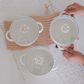 Three white ceramic bowls with matching lids, each featuring a raised white floral design in the center and a glass lid, arranged on a wooden tray over a white lace tablecloth. Two hands are holding a bowl.