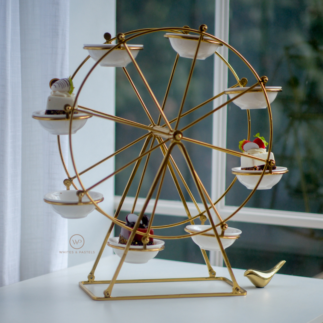 A gold metal Ferris wheel dessert stand with eight white ceramic bowls, displayed on a white table in front of a window.