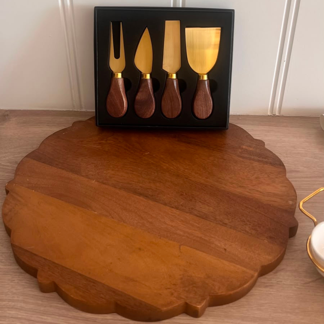 A wooden cheese knife set with gold-toned metal blades and dark brown wooden handles, displayed in a black gift box on a wooden surface with a large round wooden serving board in front.