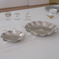 Two silver-toned scalloped ceramic or glass bowls placed on a white table runner, with printed feature icons along the top left and a small gold spoon in the background.