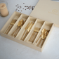 A boxed set of gold-toned flatware arranged in a beige gift box, with the matching lid placed beside it on a light surface. The set includes forks, knives, and spoons, and a candle and scattered coffee beans appear nearby as styled props.