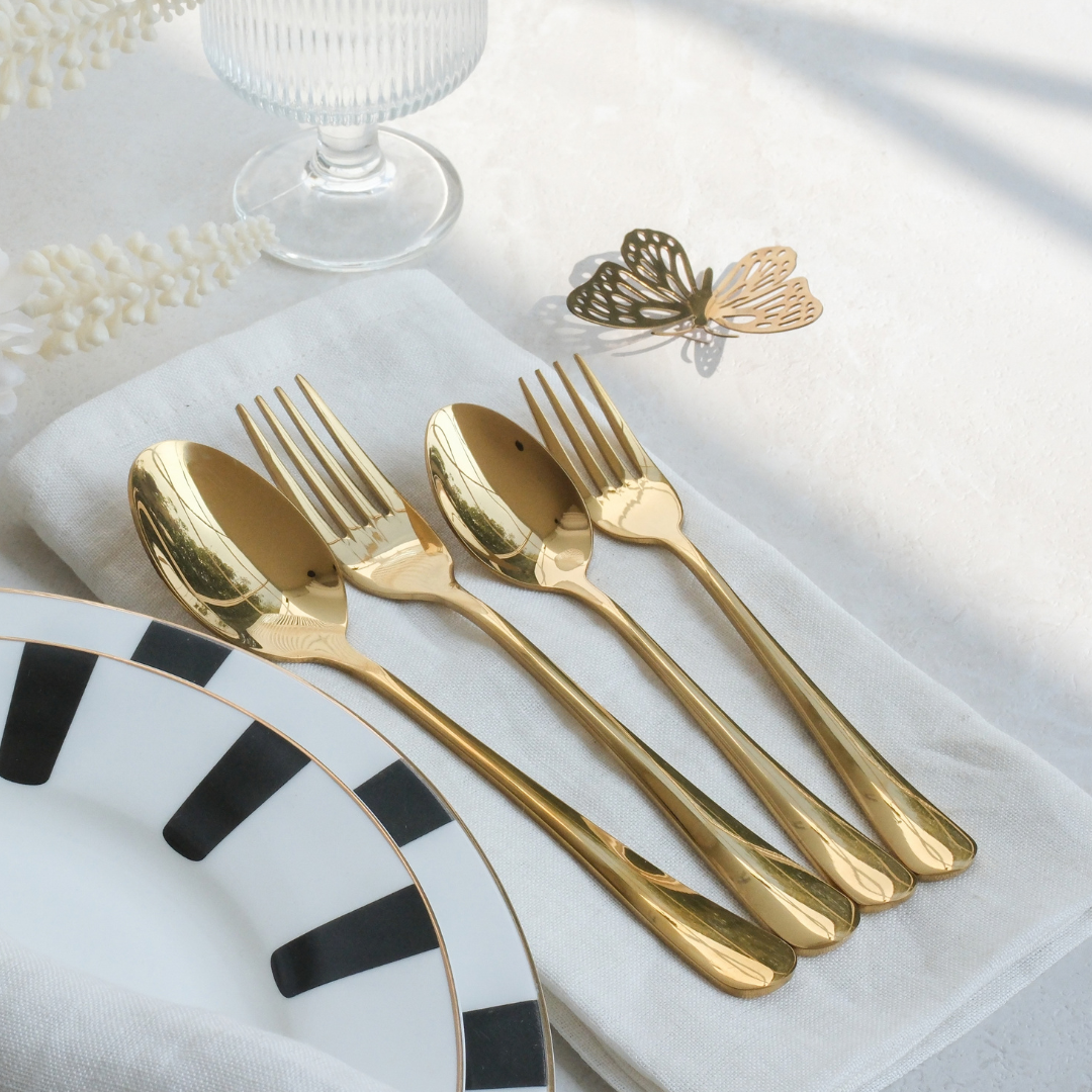 Four shiny gold flatware pieces, including two spoons and two forks, arranged on a white napkin with a black-and-white patterned plate, a glass base, and a butterfly ornament in the background.