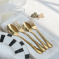 Four shiny gold flatware pieces, including two spoons and two forks, arranged on a white napkin with a black-and-white patterned plate, a glass base, and a butterfly ornament in the background.