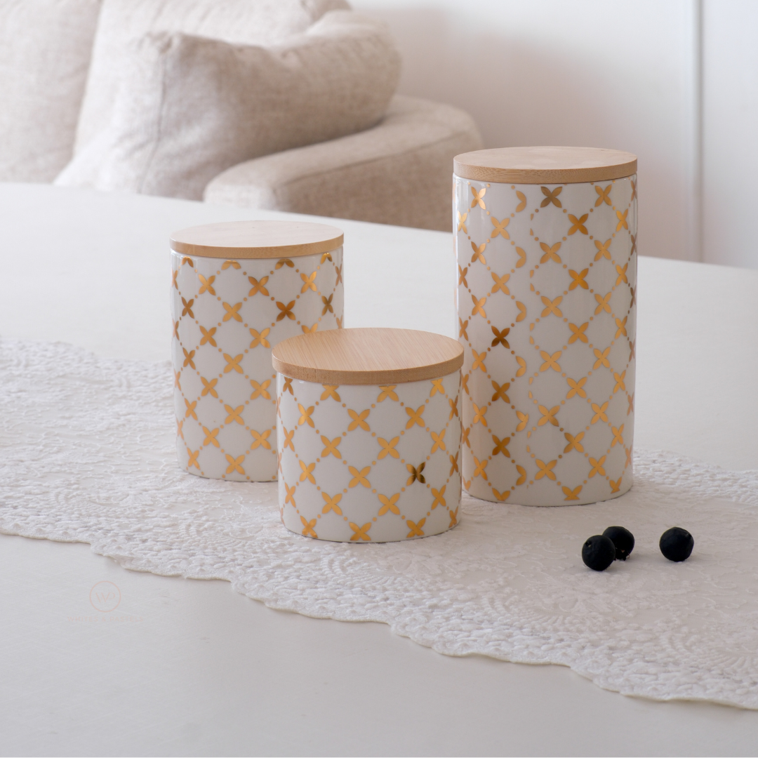 Three cylindrical storage canisters with light wood lids, featuring a white and gold geometric trellis pattern, arranged on a white lace runner in a softly lit room.