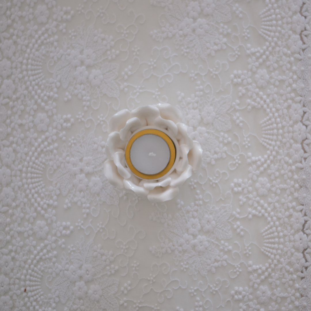 A white ceramic flower-shaped candle holder containing a tealight candle, set against a textured white lace background.