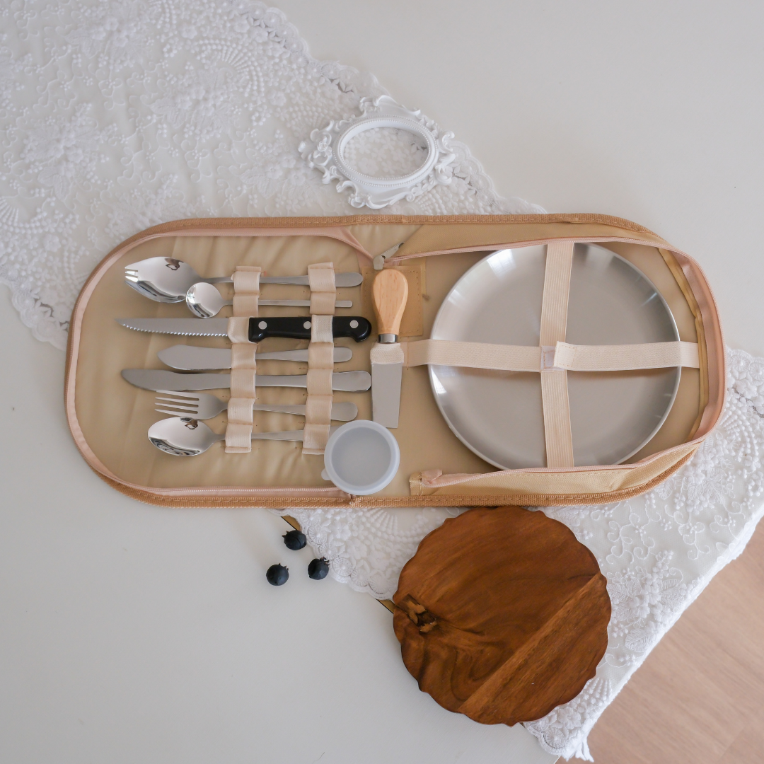 A beige picnic blanket organizer containing a silver plate, silver cutlery, a small cup, and a wooden knife, placed on a white lace runner with a wooden bowl nearby. A perfect gifting option under Rs.2000/-