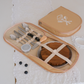 A beige picnic set organizer case shown open with stainless steel cutlery, a small cup, and a wooden board secured by cream straps, placed on a white lace cloth beside its matching closed lid.