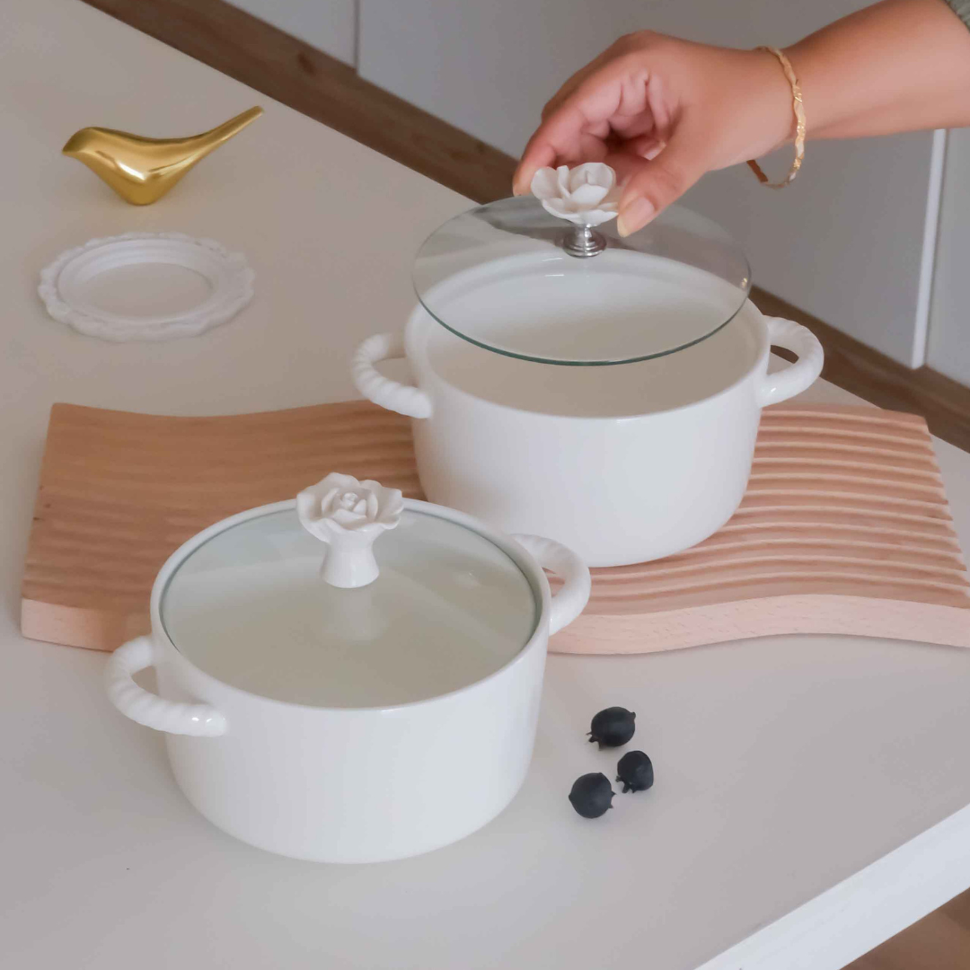 Two white ceramic pots with glass lids and white rose-shaped knob handles, displayed on a light wood cutting board on a white countertop. A hand is lifting the lid of the back pot, and a small gold bird-shaped utensil and a white plate are visible nearby.