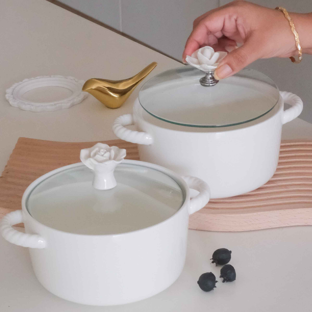 Two white ceramic pots with glass lids and decorative white rose-shaped handles, shown on a countertop with a hand lifting the lid of the larger pot. A gold bird-shaped object and a small white dish are also visible in the scene.