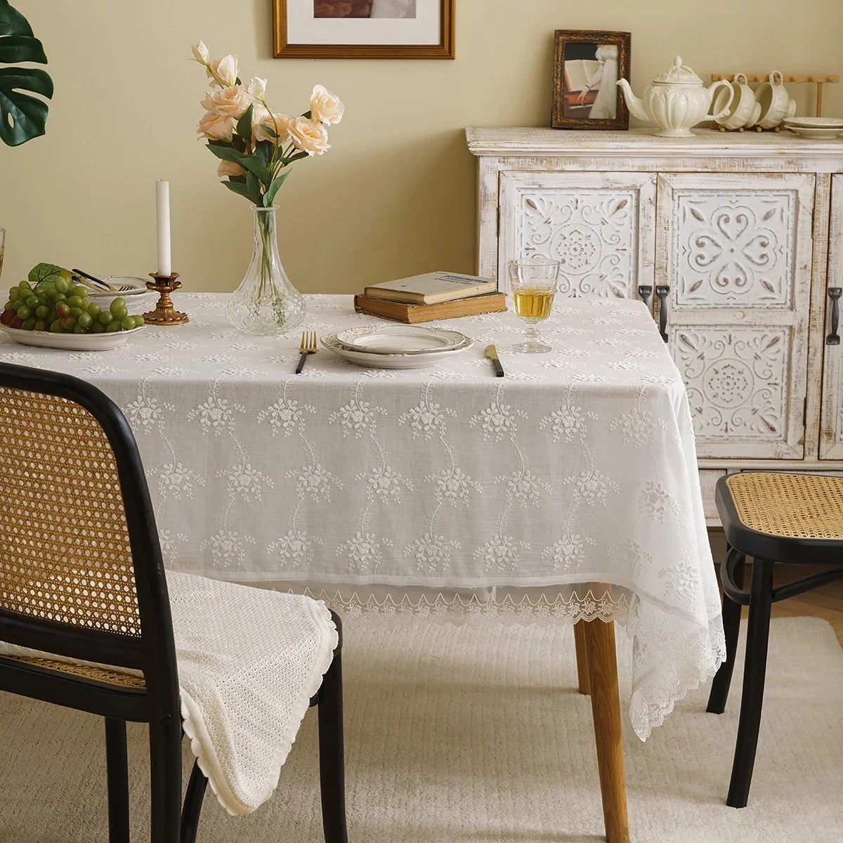 A white cotton tablecloth with European embroidery is laid out on a dining table, with intricate thread detailing visible. The table is set with cutlery and a book is placed on it, suggesting a sophisticated and elegant dining atmosphere.